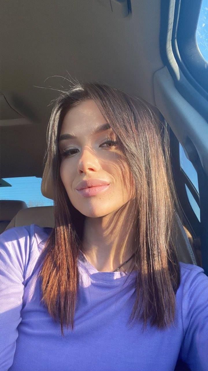 A woman in a light purple top sits in a car driver's seat, sunlight casting shadows across her face.