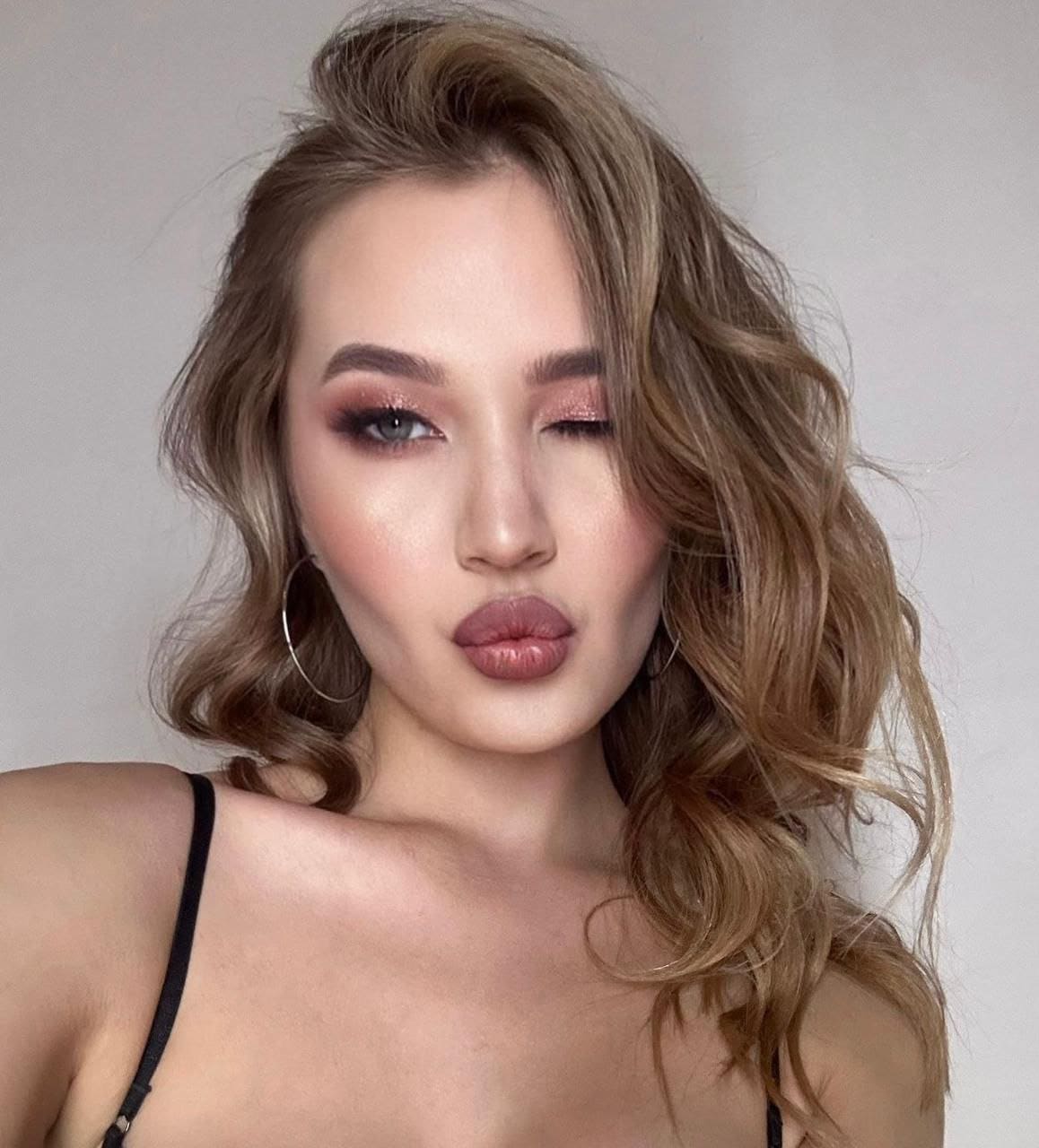 DEBBY poses with pursed lips in a black spaghetti-strap top, featuring wavy light-brown hair and pink eye makeup against a gray background.