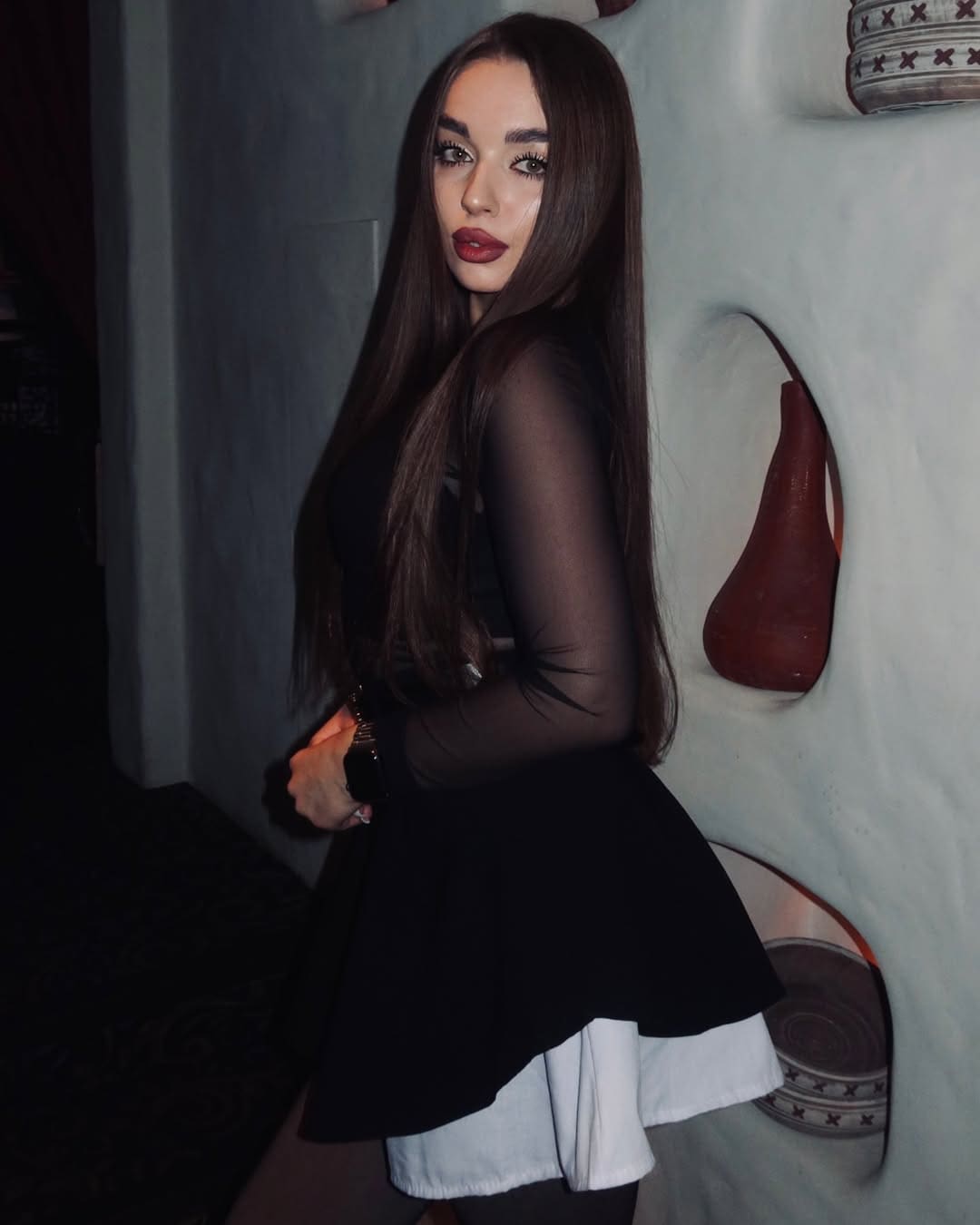 A woman in a three-quarter pose wears a black sheer-sleeved top and a layered black and white skirt against a textured wall.