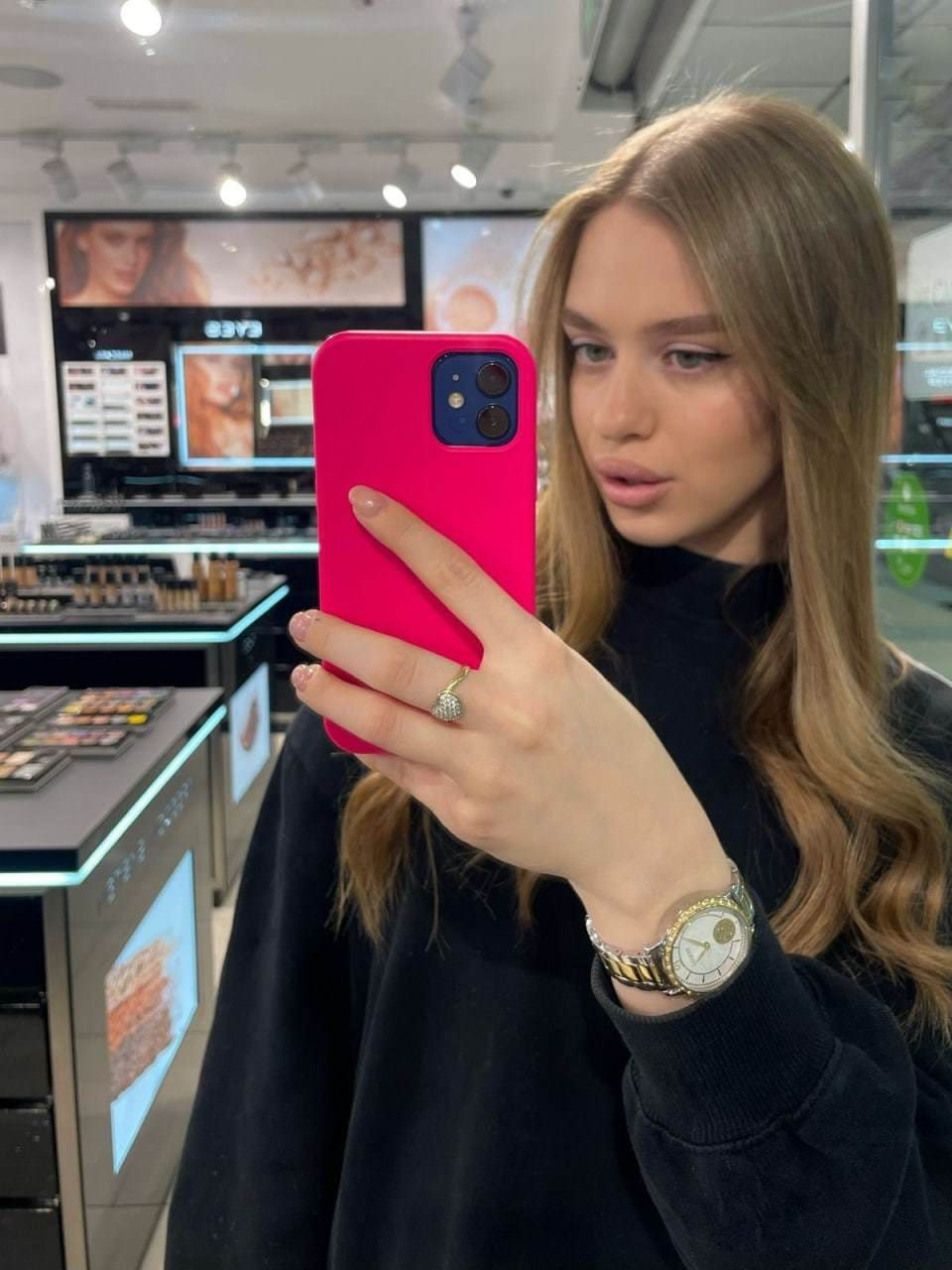 A young woman with wavy light-brown hair takes a mirror selfie in a cosmetics store, hiding her face with a pink phone while wearing a black long-sleeved top and gold ring.