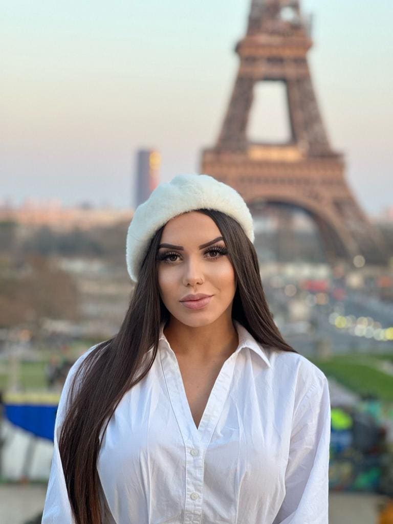 Jade stands in a white button-up shirt and fuzzy beret before a blurred iron lattice tower.