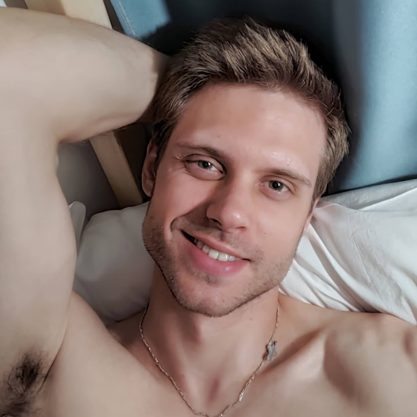A shirtless man with short brown hair lies on a white pillow, one arm raised behind his head, wearing a thin chain necklace indoors.