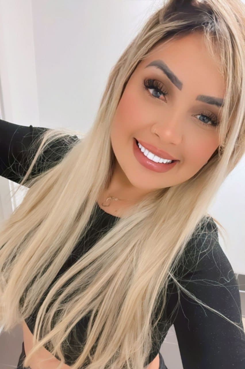 A woman in a black long-sleeved top smiles at the camera in a brightly lit indoor setting.