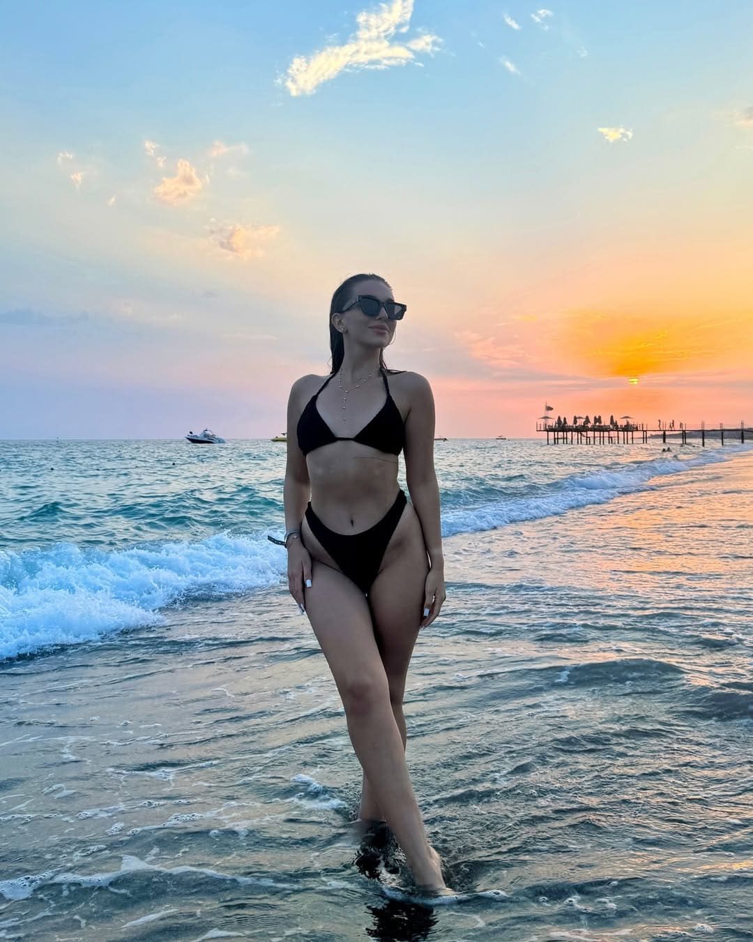 A woman in a black bikini and sunglasses stands in shallow ocean water at sunset, with waves and a distant pier behind her.