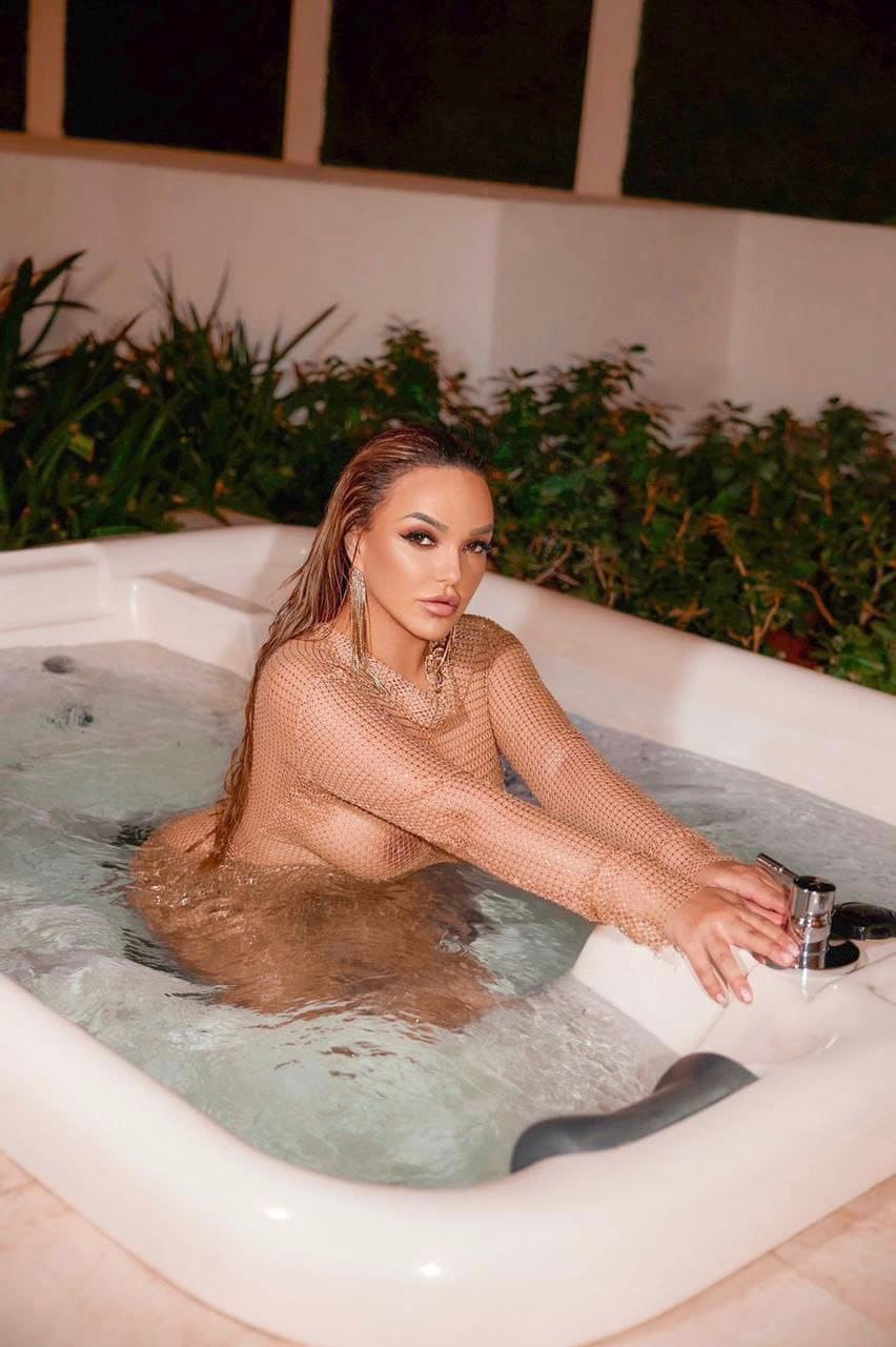 A woman with wet brown hair sits in an outdoor white bathtub wearing a sheer mesh top, with green plants and a white wall behind her.