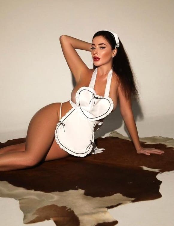 A woman in a white maid outfit with a heart-shaped bodice kneels on a cowhide rug, one hand behind her head against a light wall.