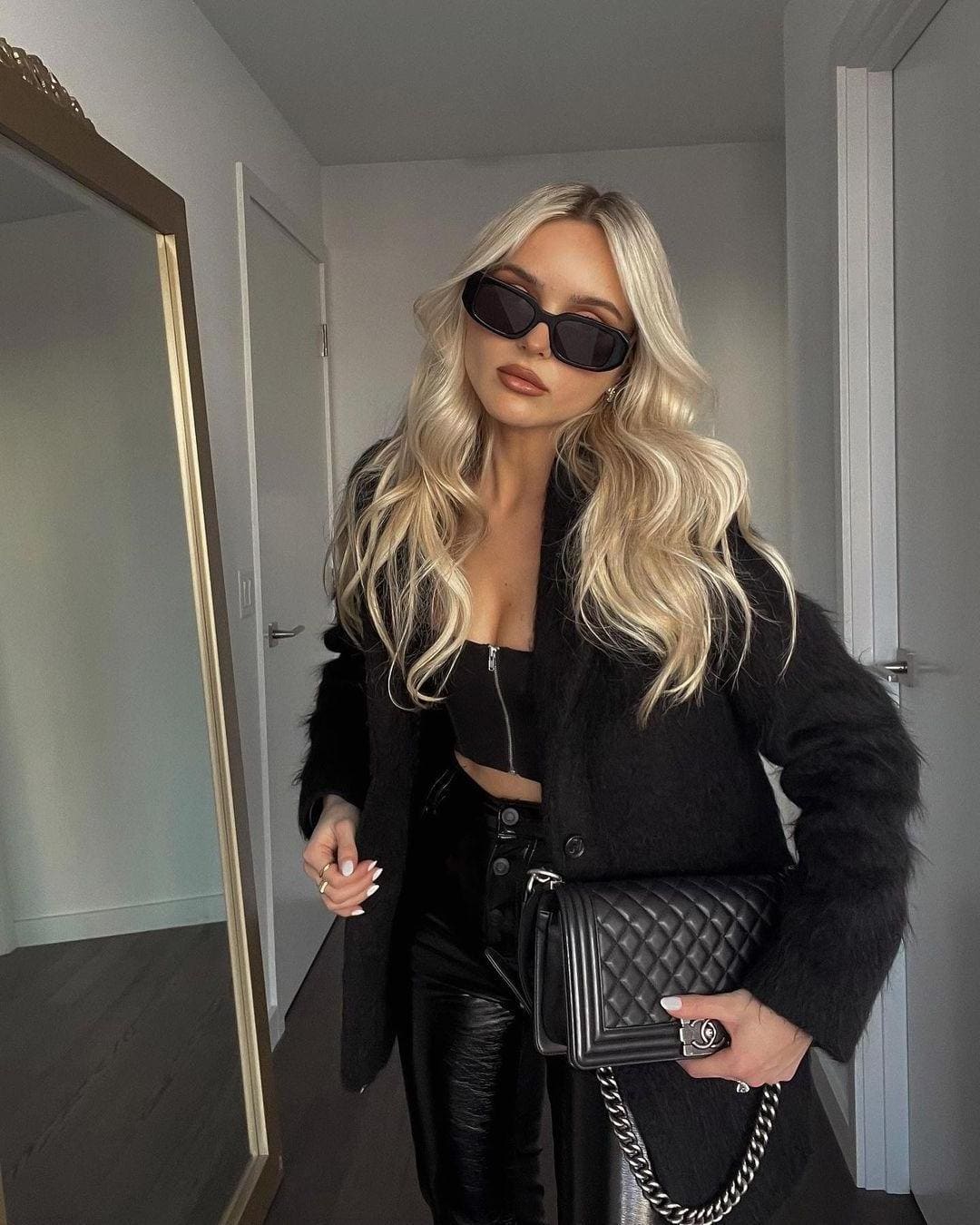 Standing in a hallway, she wears dark sunglasses, a black fuzzy coat over a crop top, leather pants, and holds a quilted chain handbag.