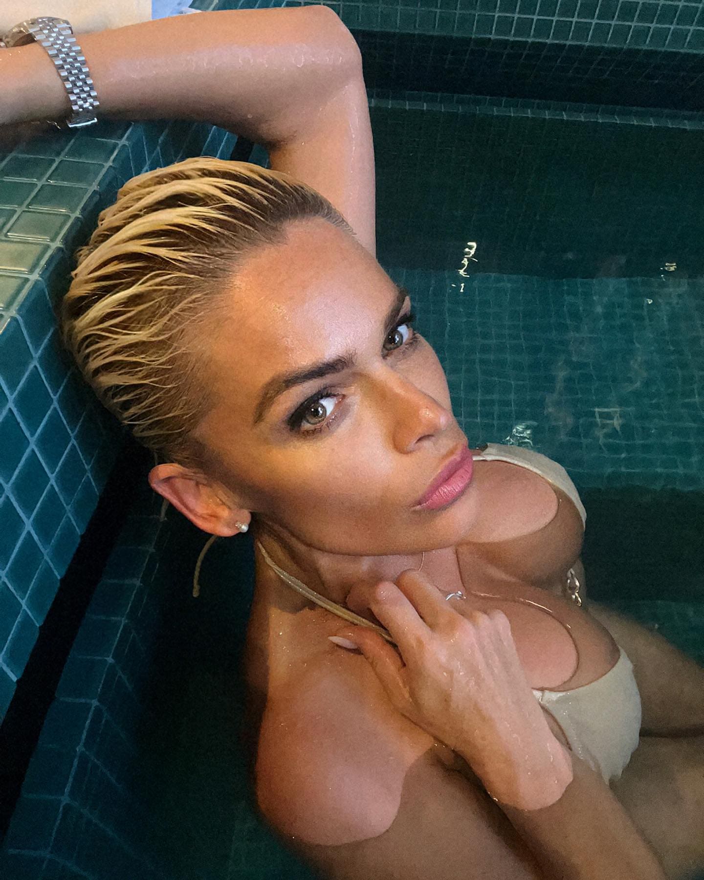 A person with wet slicked-back blonde hair is submerged in a tiled pool, wearing a light one-piece swimsuit with arms raised.