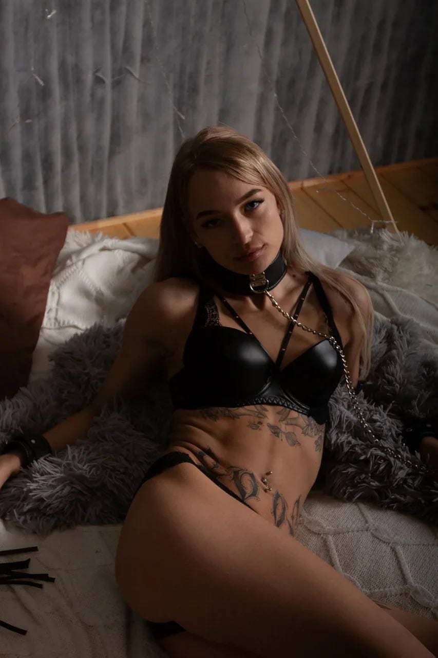 A blonde woman with midsection tattoos sits on a bed with a grey fur throw, wearing black strappy lingerie and a chain necklace.