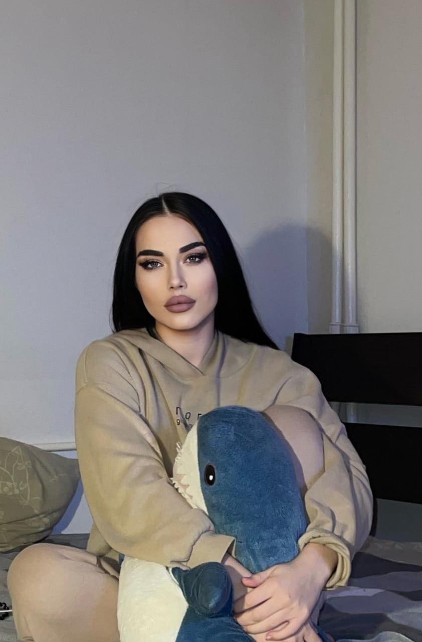 A woman sits on a bed in a beige hoodie, holding a large blue and white shark plush toy against her chest.