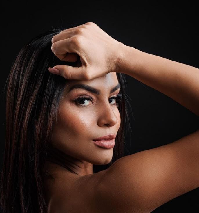 ANTONELLA poses against a black background, right hand raised to forehead, revealing bare shoulders and upper chest.
