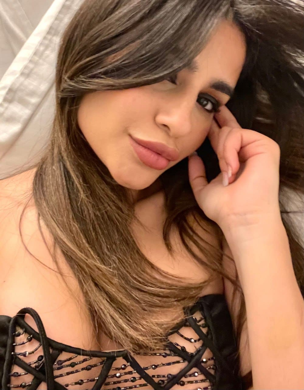 Resting her head on her hand indoors, wearing a black strappy top with beaded details, against a light-colored fabric background.