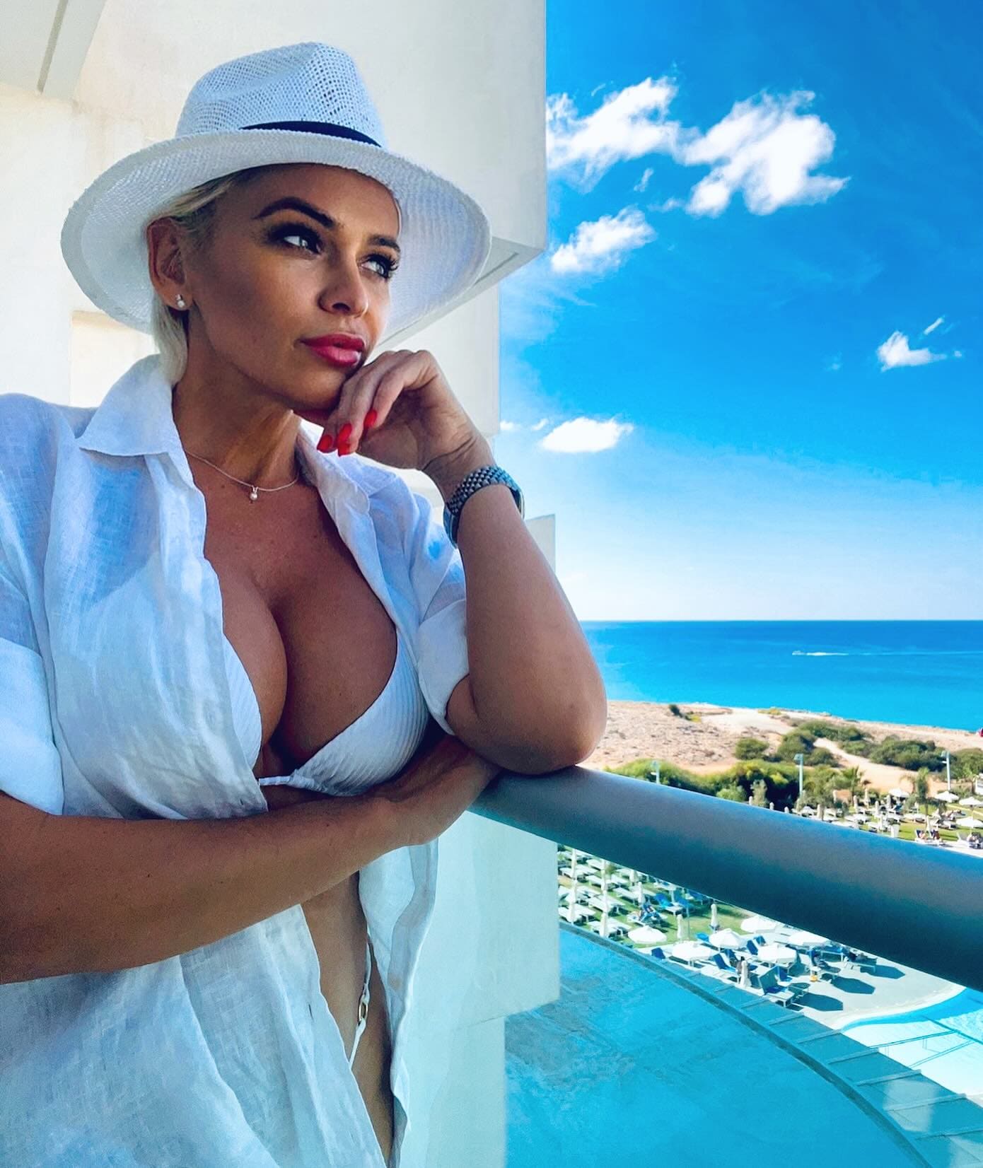 A blonde woman in a white sun hat and open shirt over a bikini leans on a balcony railing, overlooking a pool and ocean.