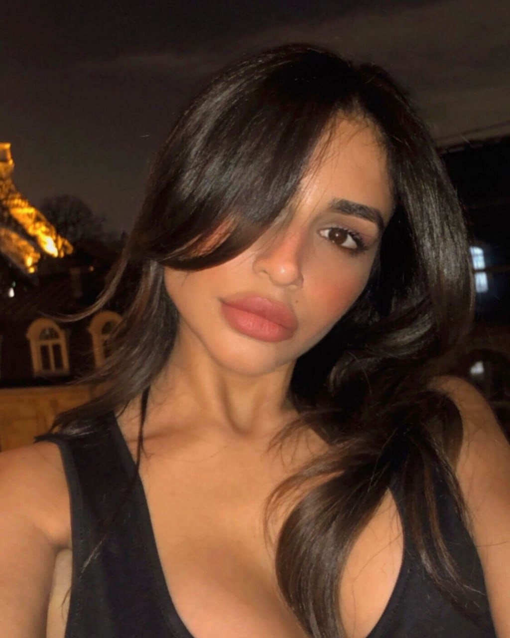 Posing at night in a black sleeveless top with a deep neckline, hair covering one eye, with a lit building in the background.