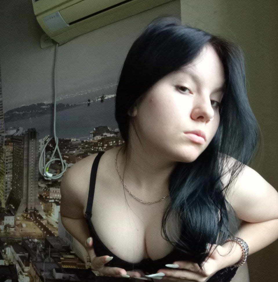 Olesya poses indoors in a black low-cut top and necklace, hands near her chest, against a cityscape wallpaper.