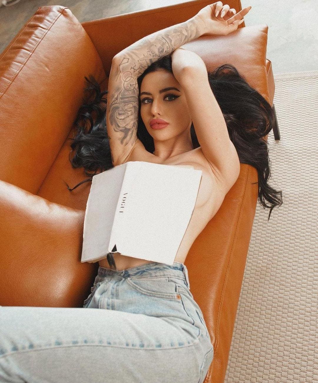 A woman with a tattooed arm lies in a tan armchair, wearing a white strapless top and jeans, holding an open book.