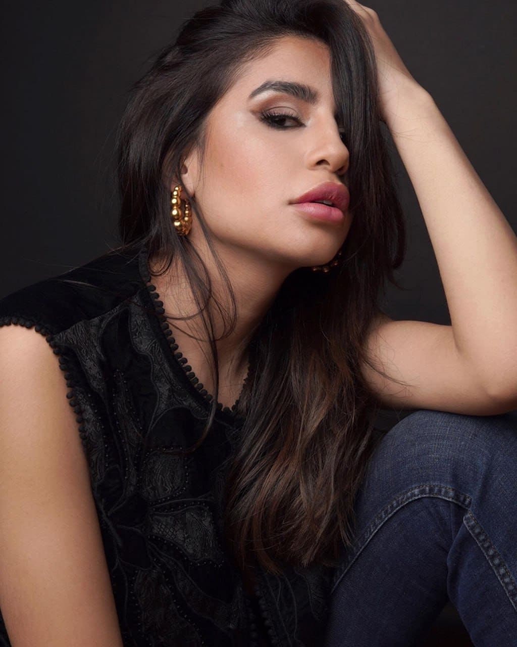 Sitting against a dark background in a black lace top and dark jeans, one hand raised to her head, wearing gold hoop earrings.