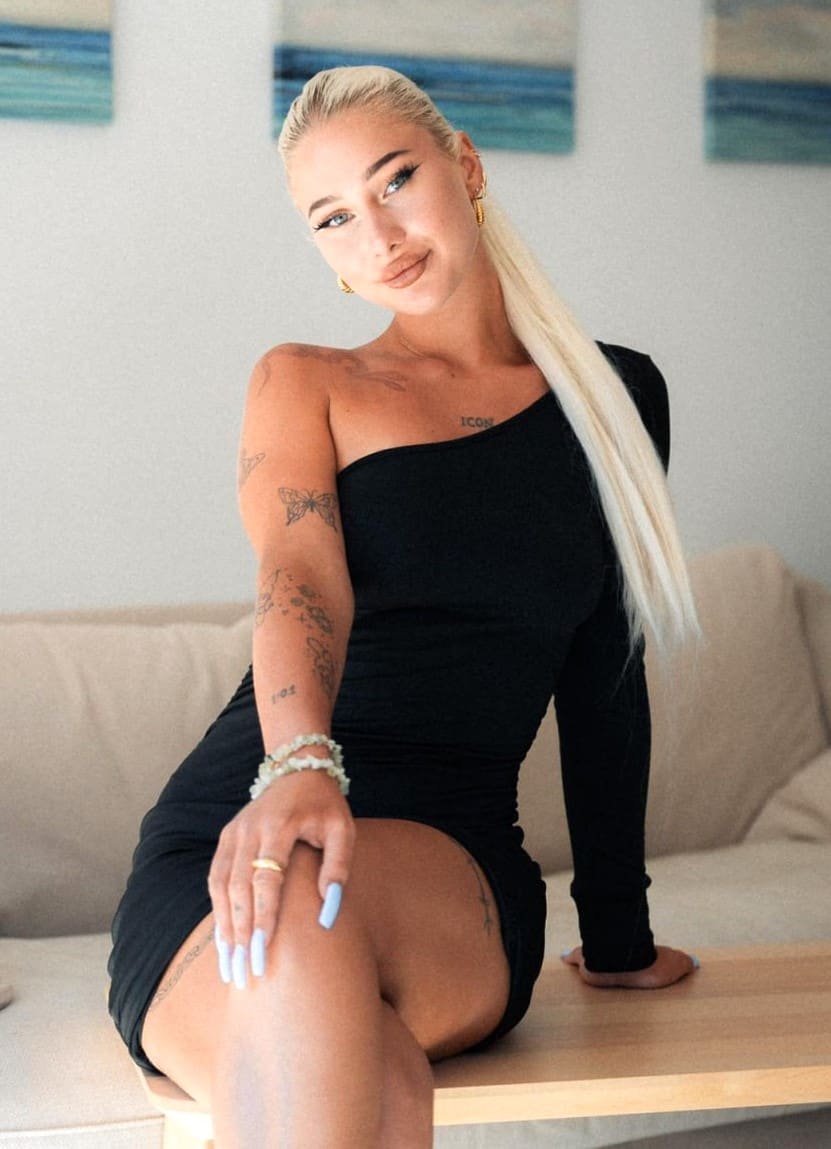 A blonde woman with a high ponytail and arm tattoos sits on a wooden table wearing a black one-shoulder mini dress, with a beige sofa and wall art.