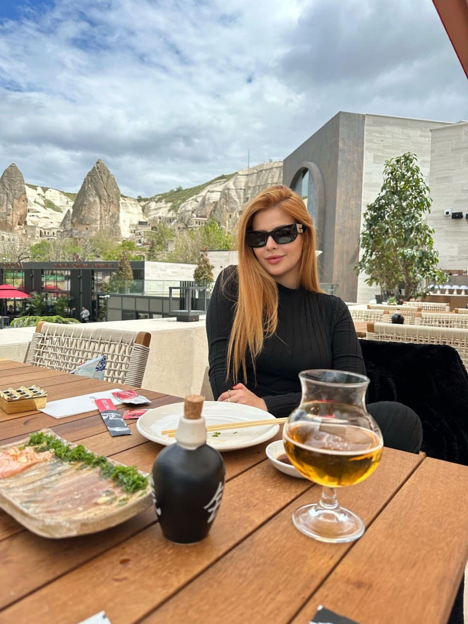 A woman in sunglasses and a black top sits at an outdoor table with food and a drink, stone arches and hills in the background.