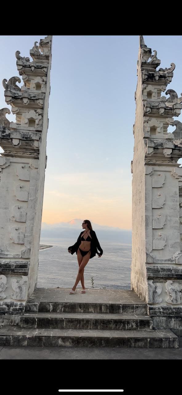 Woman in a black bikini and open cardigan stands on stone steps between white temple gateways, with water and mountains visible at dusk.