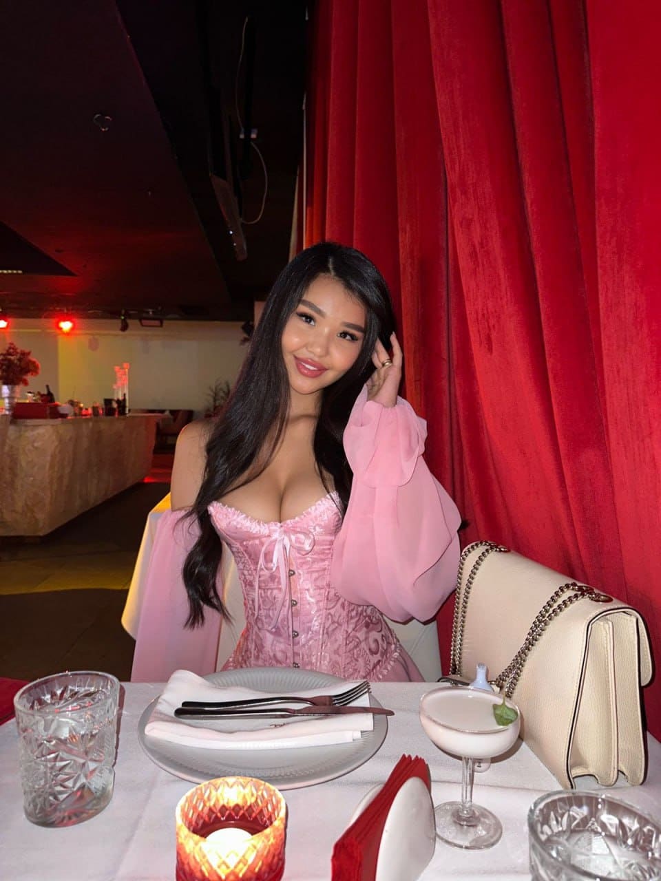 Seated at a restaurant table, a woman in a pink off-the-shoulder corset top touches her hair near a red curtain.