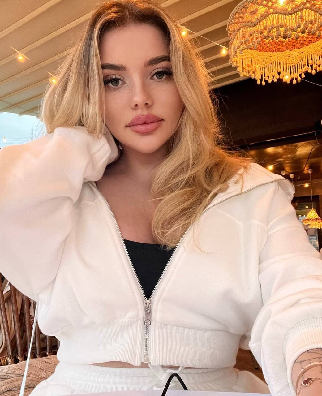 Lesya in a white cropped hoodie and black top poses indoors near a chandelier, with string lights visible in the background.