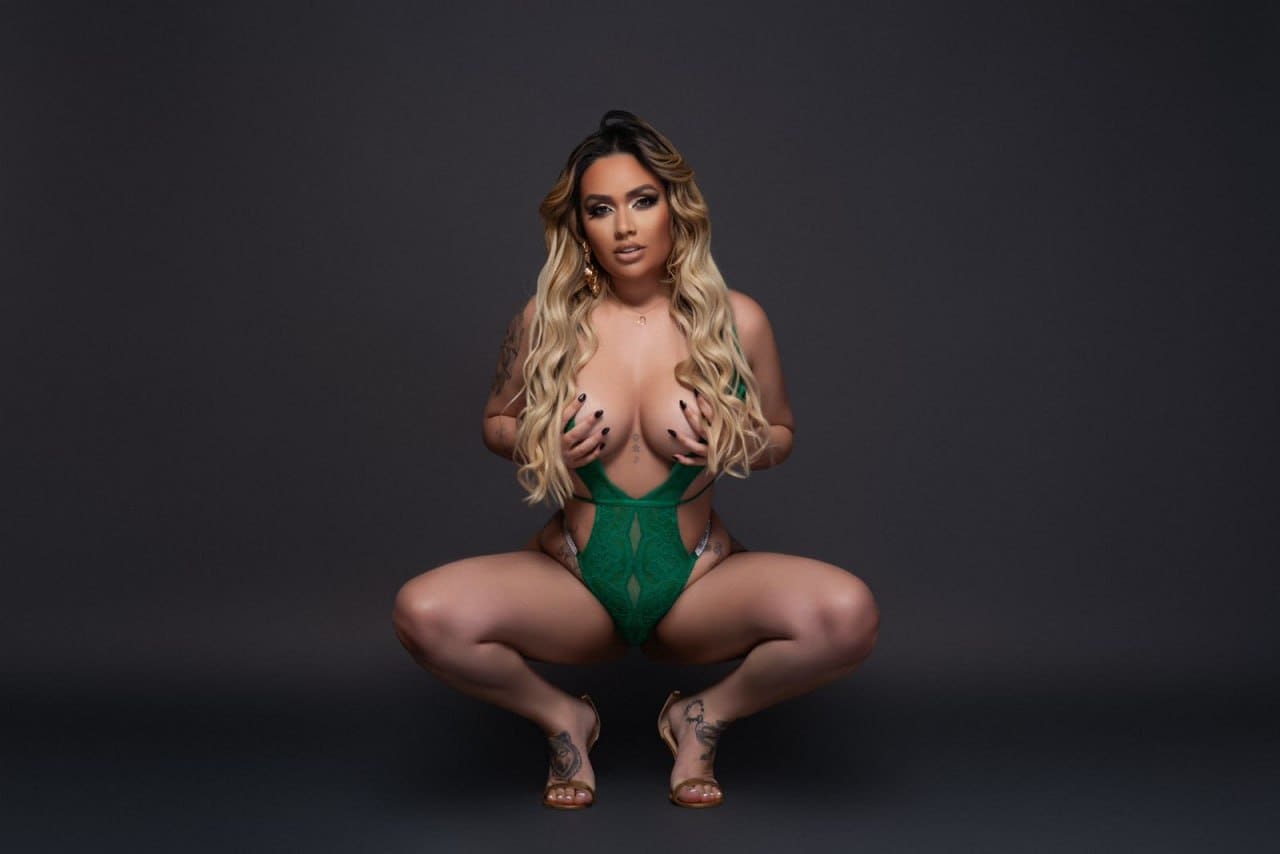 A woman crouches in a green lace bodysuit and strappy sandals against a dark studio backdrop.