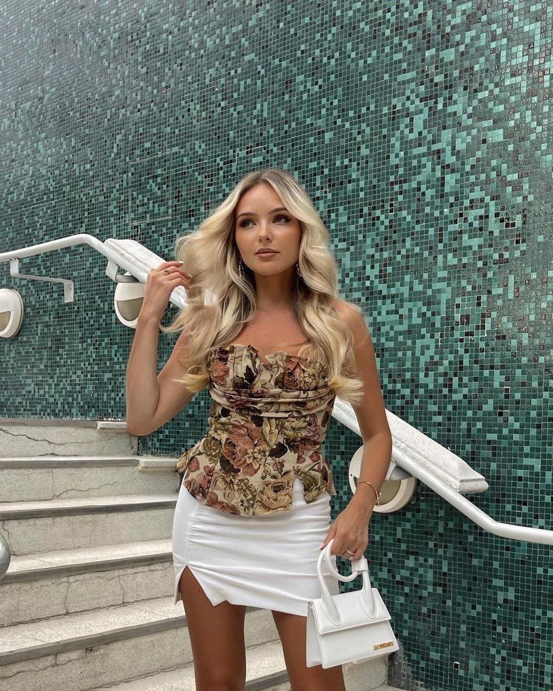 Celeste stands on stone steps before a green mosaic wall, wearing a floral peplum top and white mini skirt, holding a white handbag.