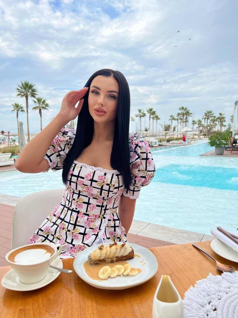 Sitting at a poolside table, she wears a floral grid dress, touching her hair near coffee and dessert under palm trees.