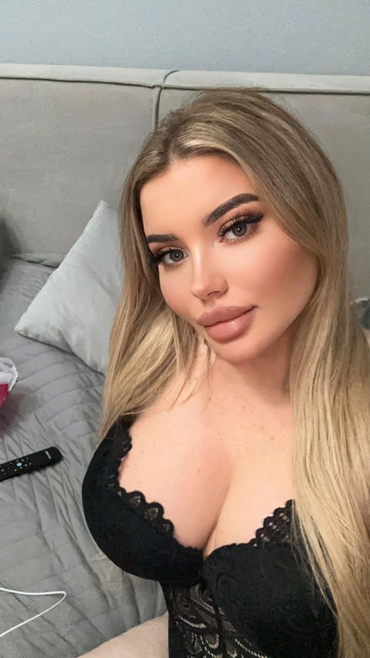 A woman in a black lace bustier sits on a grey bed facing the camera, with a remote control and pillow visible behind her.