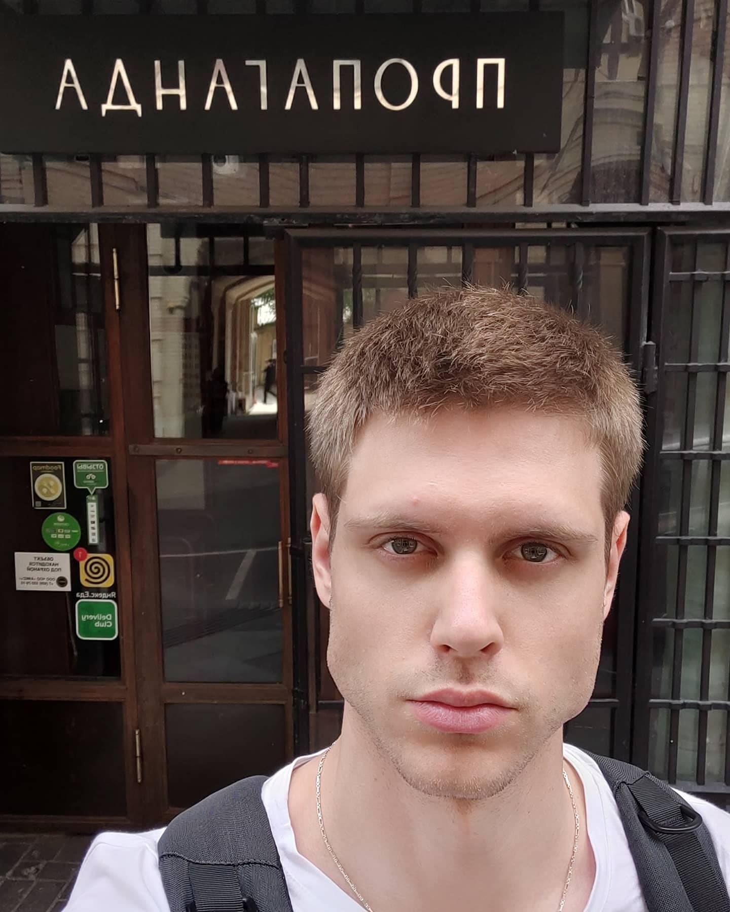A young man in a white t-shirt and dark backpack stands before a storefront with a black sign, wearing a thin chain necklace.