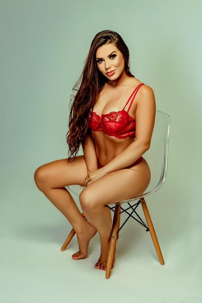 Sitting on a clear chair with hands on lap, she wears a red lace bra and underwear against a light green background.