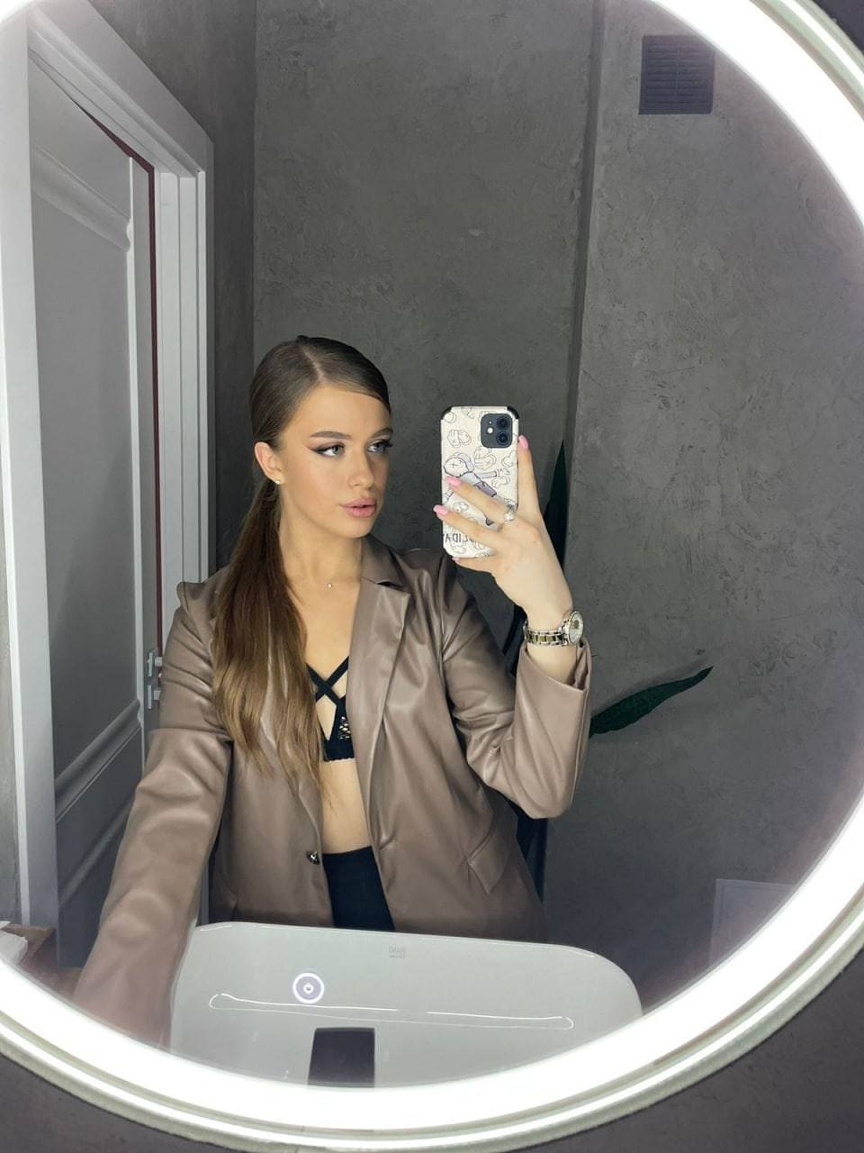A woman with a side ponytail takes a mirror selfie in an open satin blazer over a black strappy top, standing in a room with gray walls and a white door.