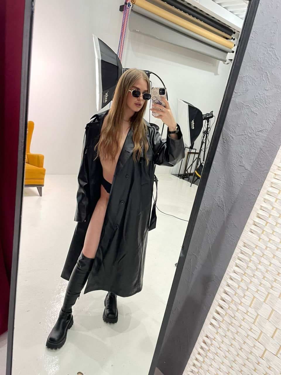 A blonde woman with sunglasses takes a studio mirror selfie wearing an open black leather trench coat over a black bra and high black boots.