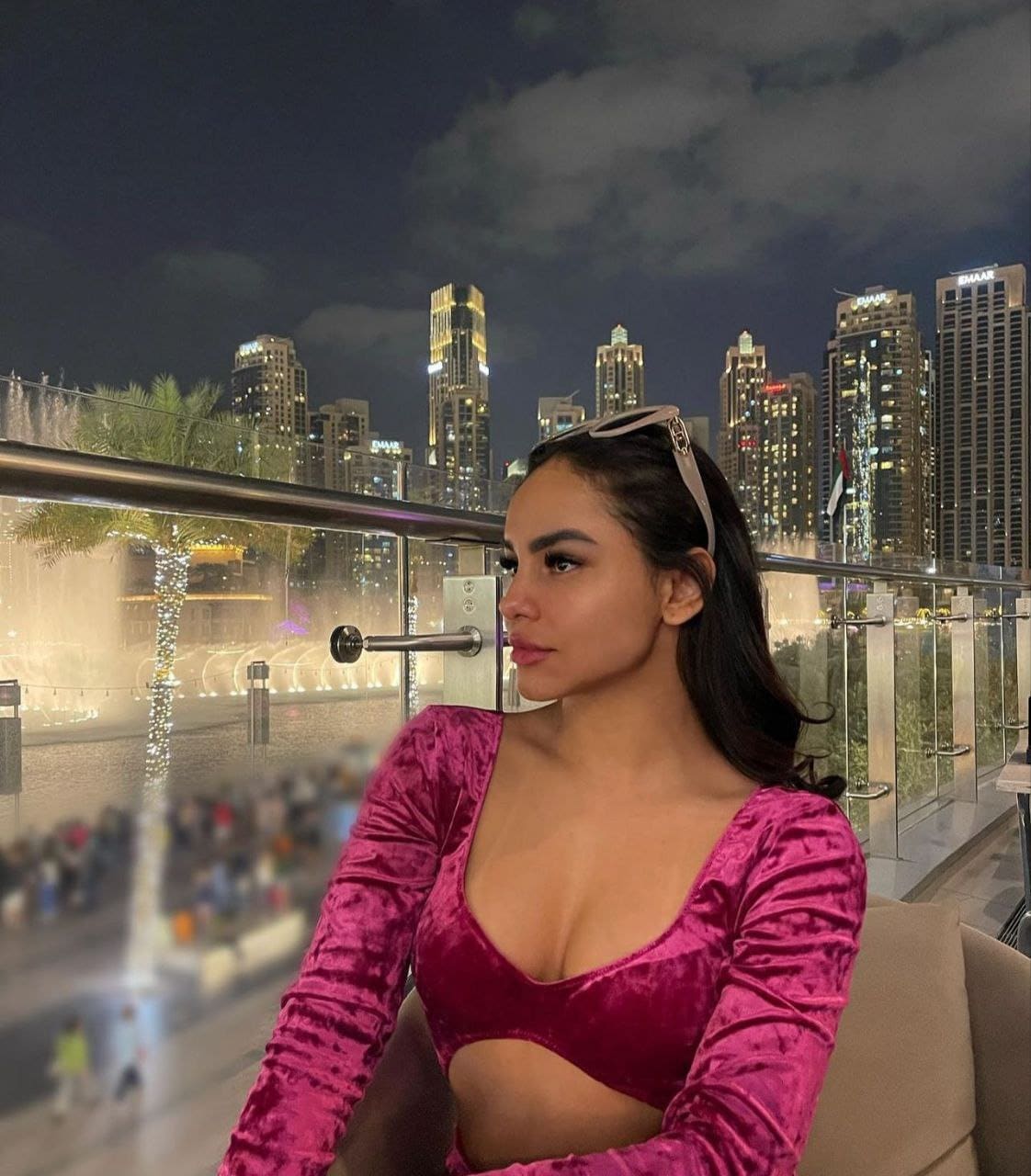 Woman in profile wearing a magenta velvet crop top and sunglasses on head, seated before a night city skyline and fountain.