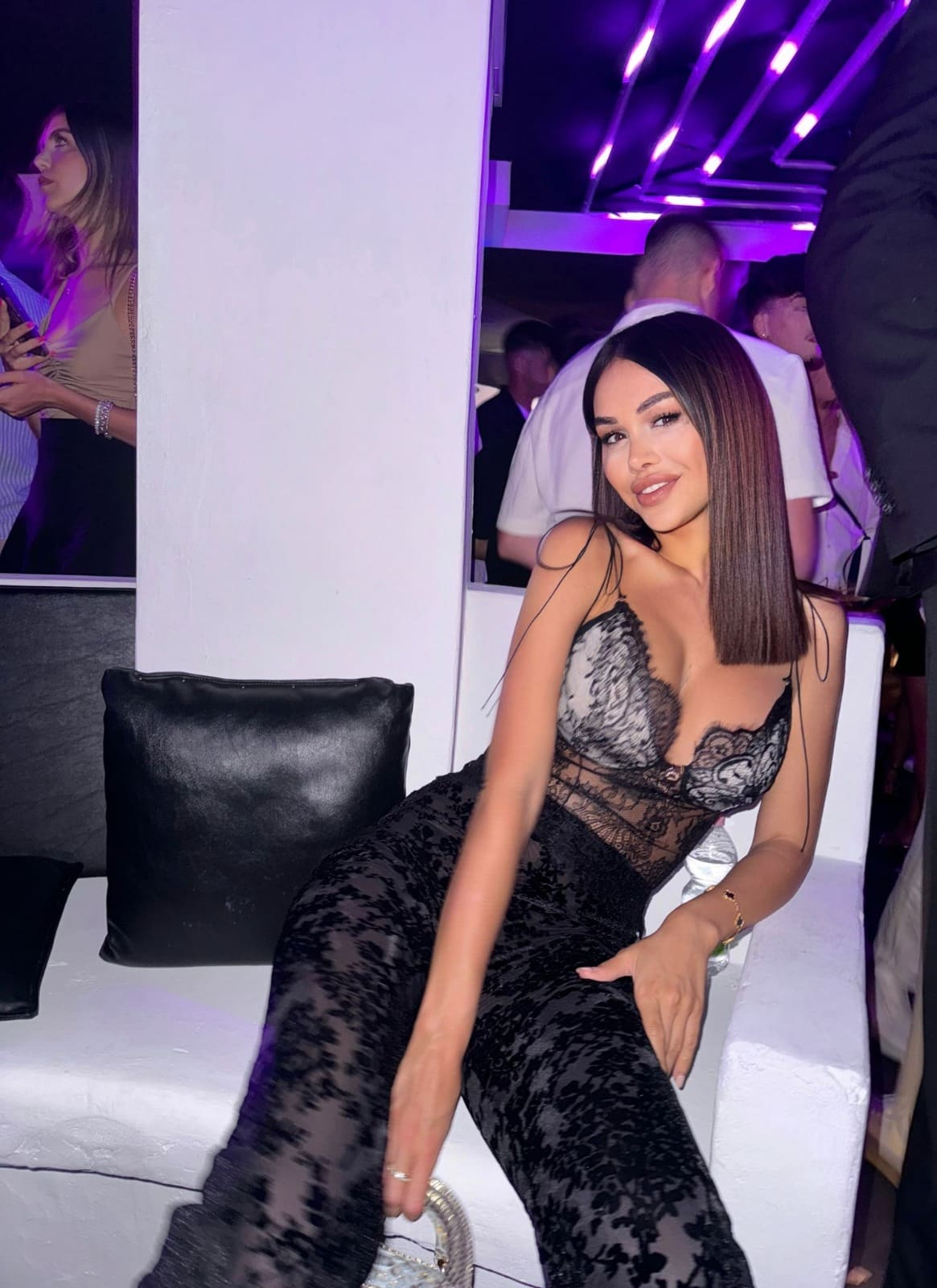 A woman in a black lace bodysuit sits on a white bench under purple lights, one hand resting on her thigh.