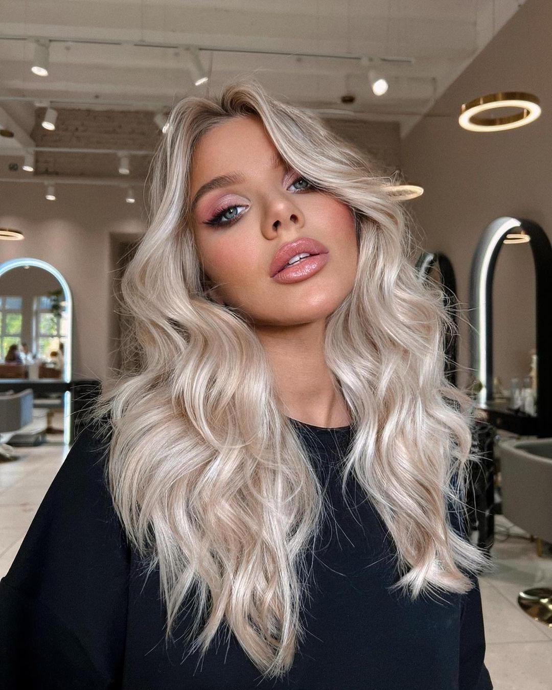 A woman with platinum blonde hair and pink eyeshadow poses in a black top inside a salon featuring arched mirrors and track lighting.