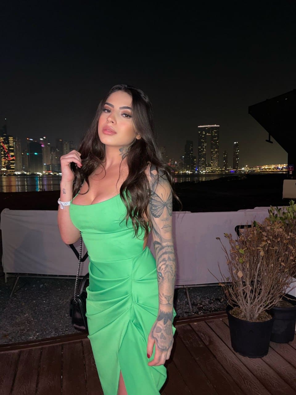 A woman with a tattooed arm stands on a wooden deck at night in a strapless green dress with a slit, city skyline behind.
