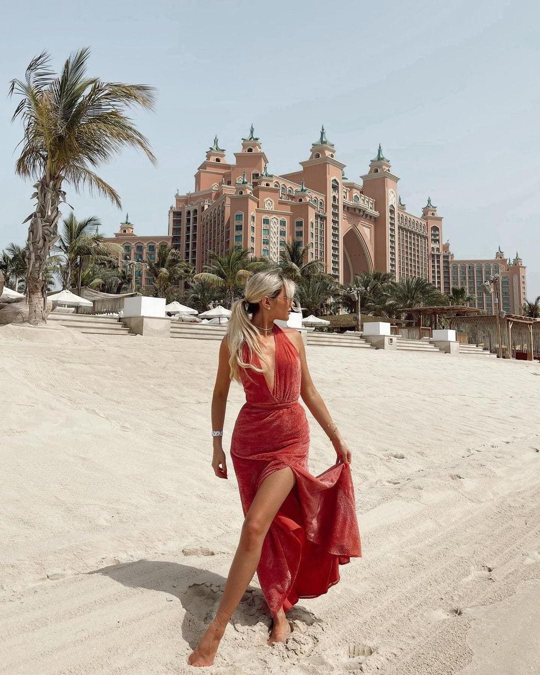 Barefoot on a sandy beach, a blonde woman in a red halter dress with a high slit holds the hem, with a pink resort and palms behind.