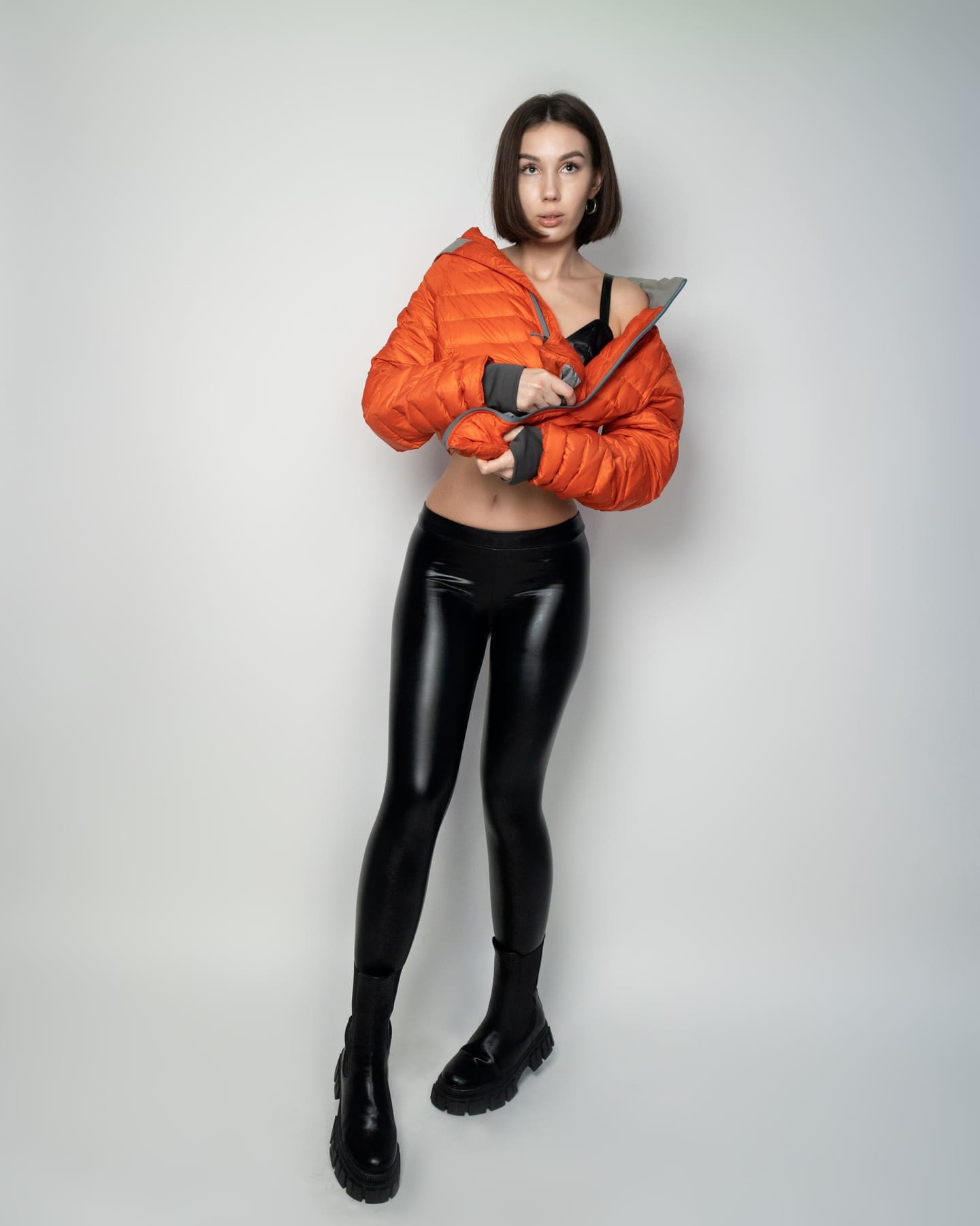 A woman with a short brown bob wears an open orange puffer jacket over a black bra top and glossy leggings, holding the jacket at her waist.