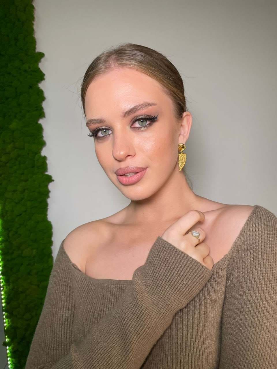 Amilina wears a brown off-the-shoulder ribbed sweater and gold earrings, posing with a hand near her collarbone against a white wall and green moss panel.
