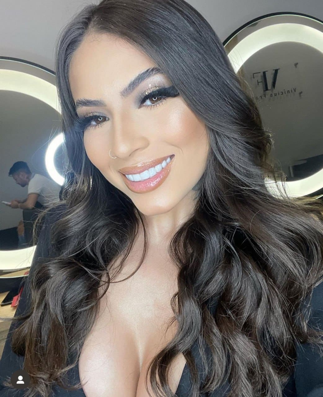 Loren smiles in a close-up selfie, wearing a black top with a deep neckline, in front of a round illuminated mirror.