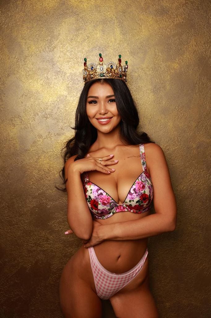Janna wears a jeweled crown, floral bra, and pink gingham briefs, posing against a textured gold backdrop.