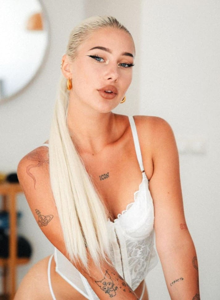 Elina with a platinum blonde side ponytail wears a white lace bodysuit indoors, posing against a plain wall with a round mirror in the background.