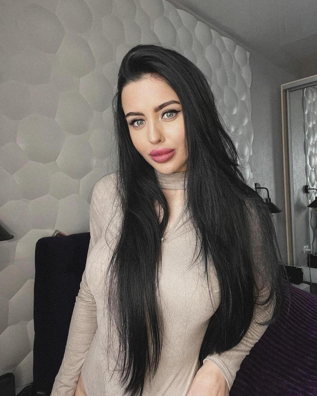 A woman in a beige long-sleeved top poses in front of a textured wall, with long dark hair visible.