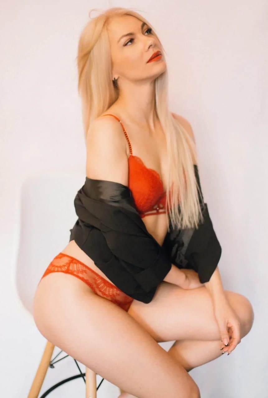 A blonde woman sits on a wooden stool in orange lace lingerie and a black satin robe, positioned against a plain white background.