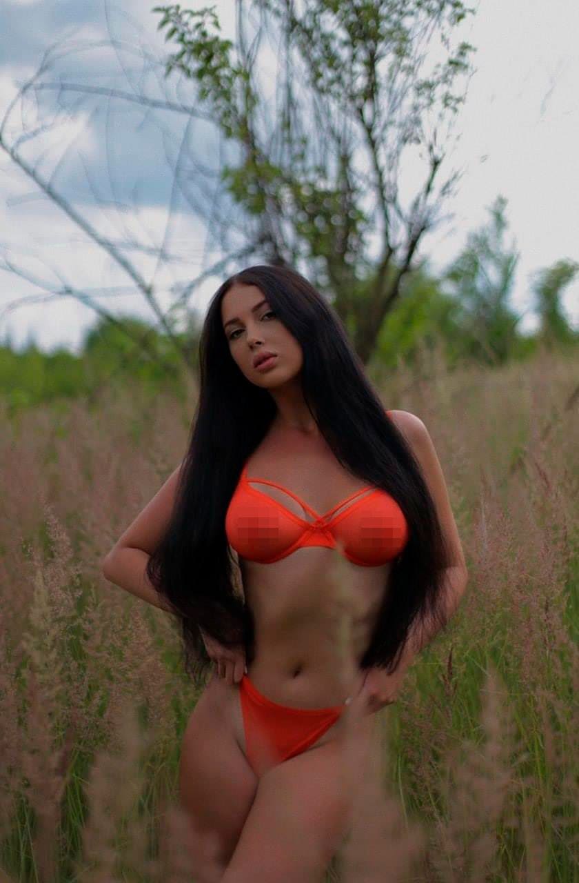 A woman in an orange bikini stands with hands on hips in tall grass under an overcast sky.