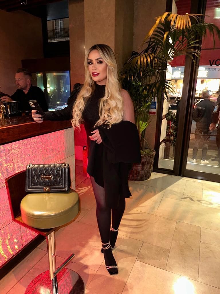 A woman in a black off-shoulder dress and tights stands in a lobby next to a bar counter and handbag.