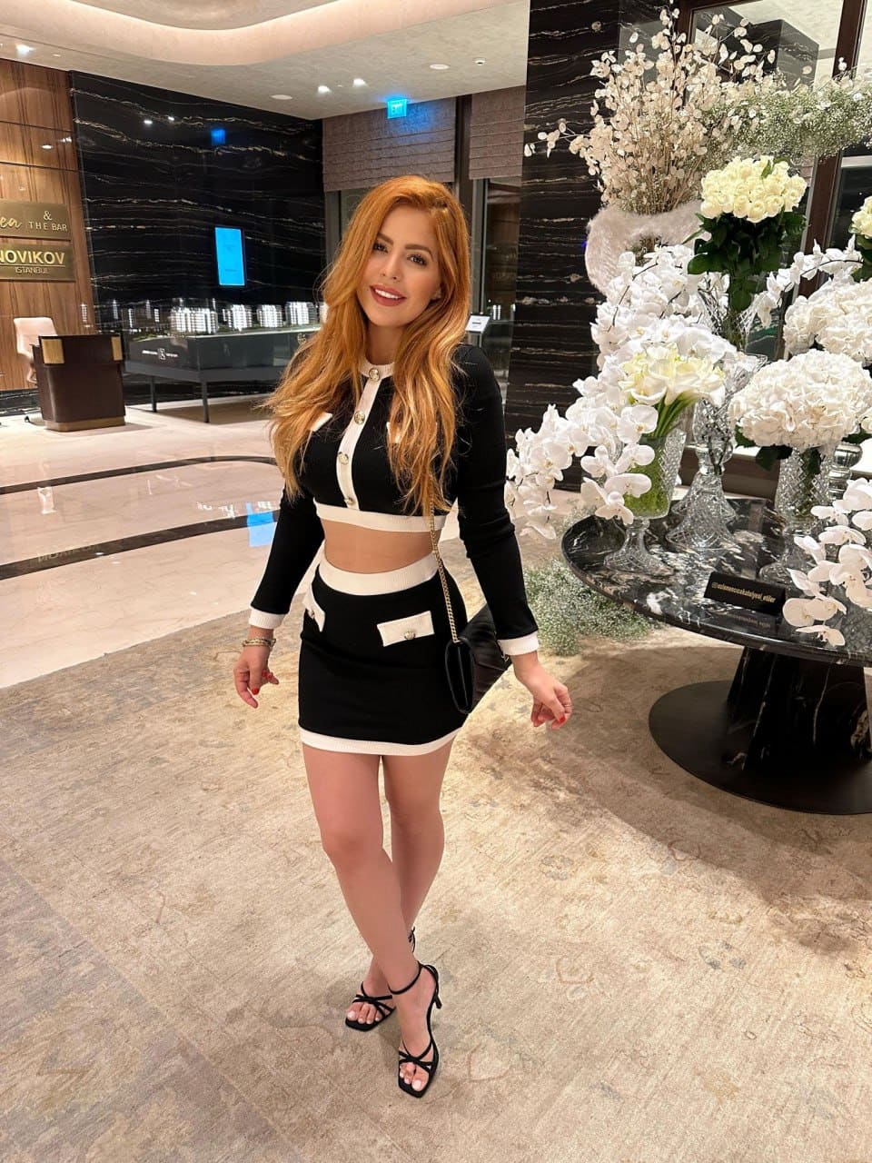 A woman in a black and white two-piece outfit and strappy heels stands in a hotel lobby beside a table with white flowers.