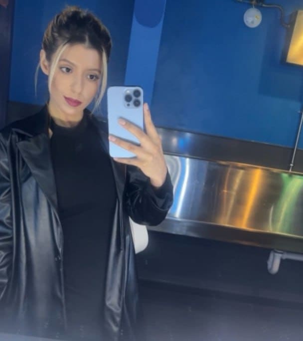 Mirror selfie of a woman in a black turtleneck and leather jacket holding a blue phone near a metallic counter.
